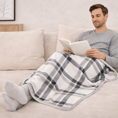 Man sitting on a couch with a grey plaid blanket draped over his legs, reading a book in a neutral living room setting